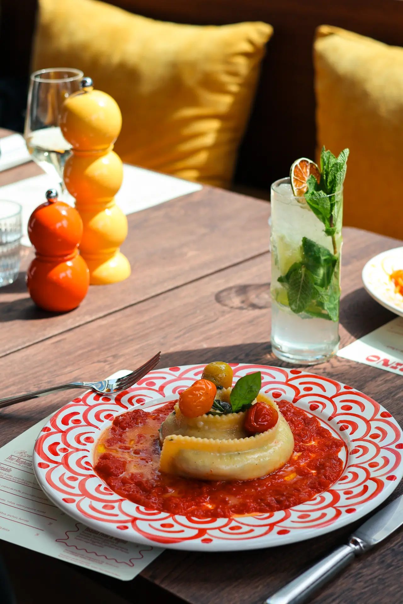 Filled ravioli on a hand-painted plate, served with tomato sauce, basil and cherry tomatoes, alongside a refreshing cocktail